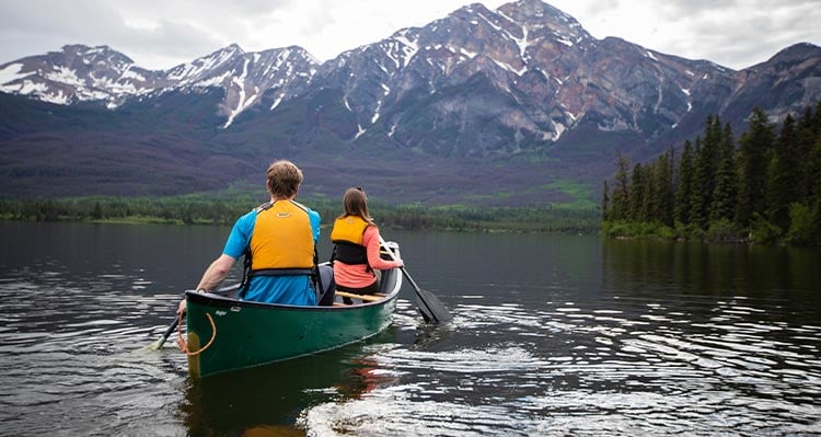 Two people canoe along a calm lake below forested mountains.