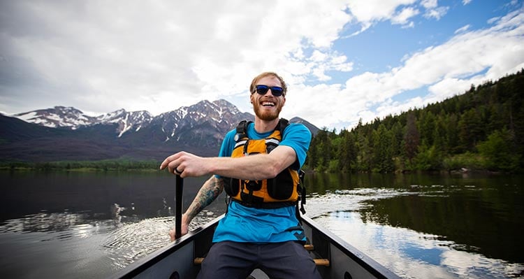 A person paddles in a canoe on a calm lake below mountains.