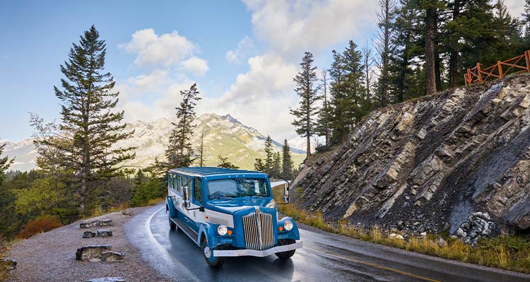 A blue vintage touring-automobile rounds a corner on a road below a small cliffside.