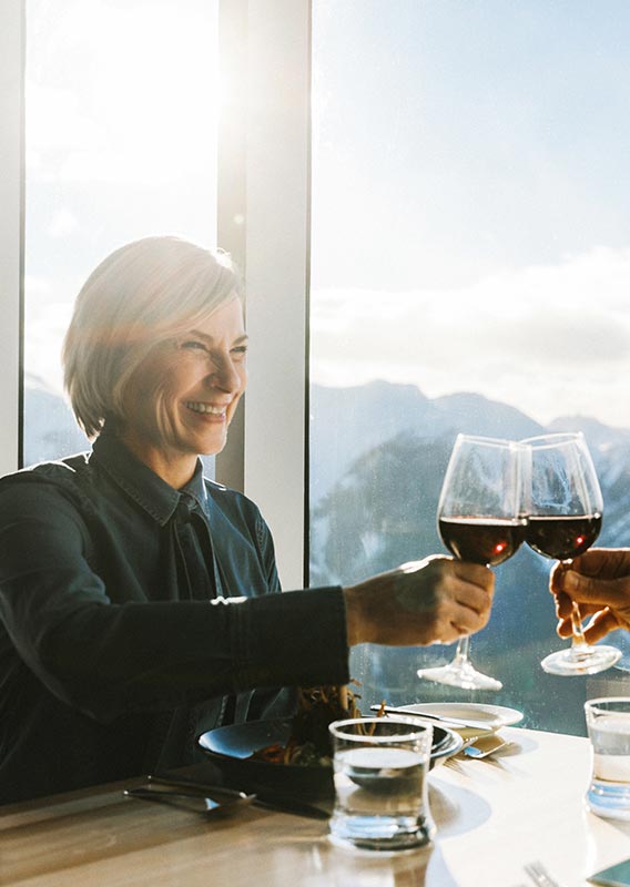 A couple raise wine glasses in a cheers at a windowside table overlooking mountains.