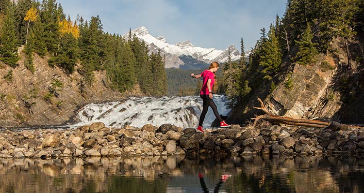 A hiker walks along rocks across a river in front of a small rushing waterfall.