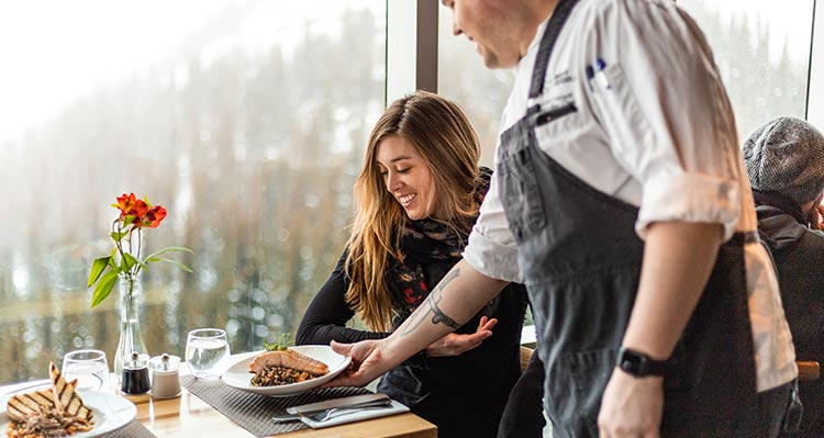 A chef brings a plate of dinner to a woman sitting at a window-side table.