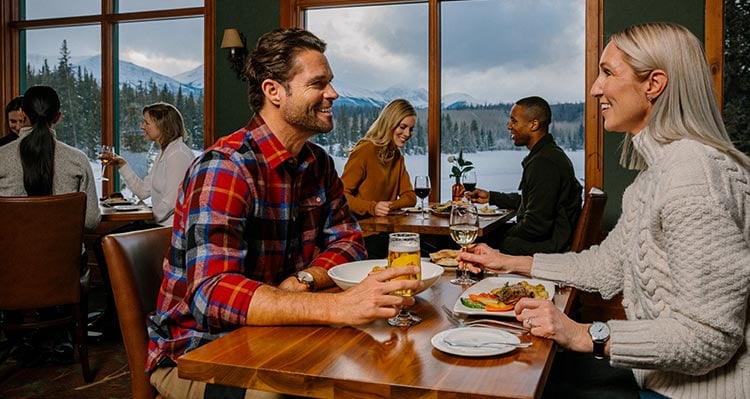 A group of friends sit at a dinner table next to a large window overlooking a winter landscape.