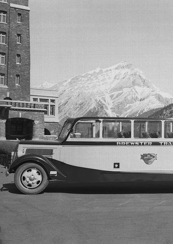 A historic photo of a tour bus parked in front of a large stone building with mountains in the back.