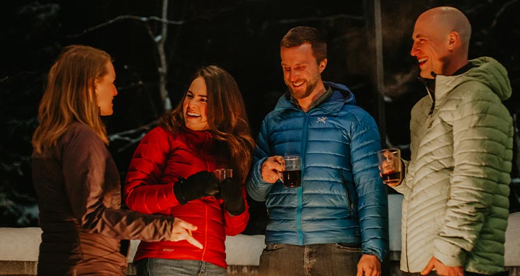 Four people stand outside by the glow of a campfire.