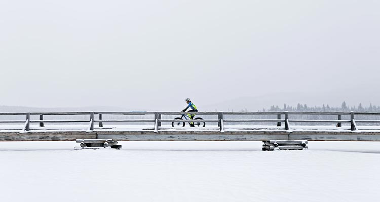 A cyclist rides across a small wooden bridge above a frozen lake.
