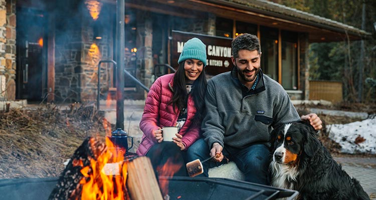 Two people sit with a dog at a campfire.