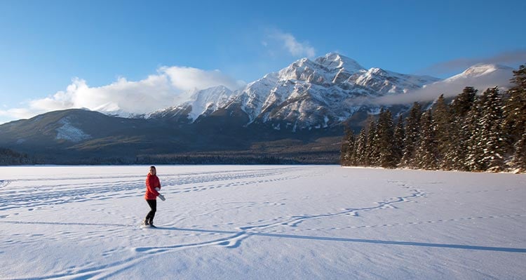 A person walks on frozen lake below snow-covered mountains.