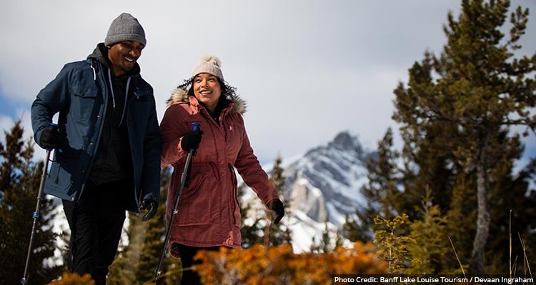 Two people hike among shrubs and trees on a trail in the winter.