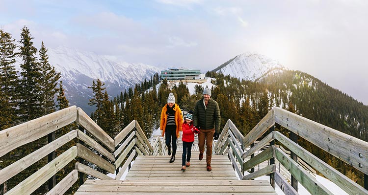 A family walks on a mountaintop boardwalk above snow covered tree slopes.