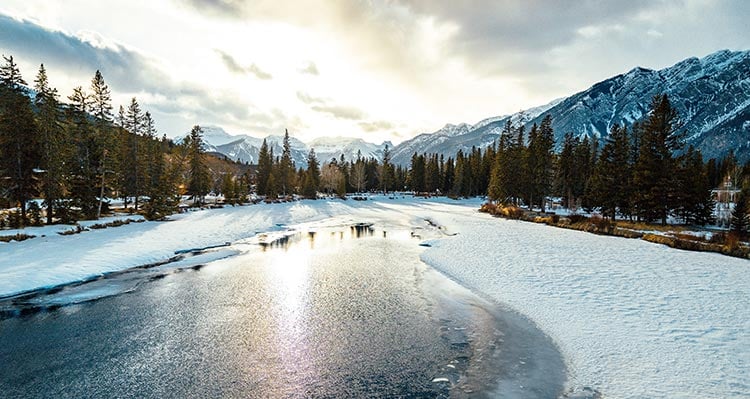 A view down a partially frozen river between forested banks below snow-covered mountains.