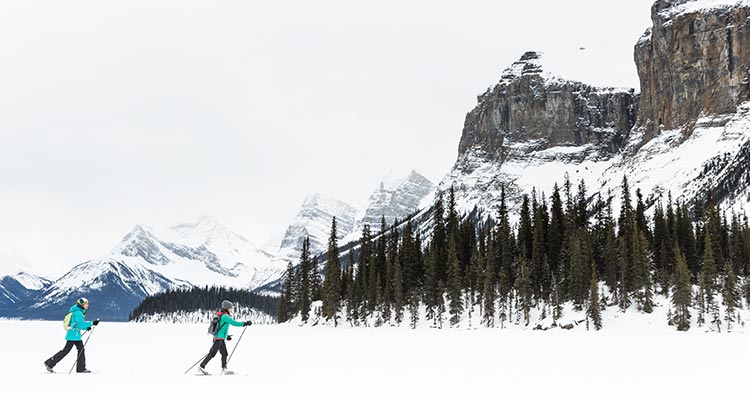 Two people cross country ski across a frozen lake below mountains.