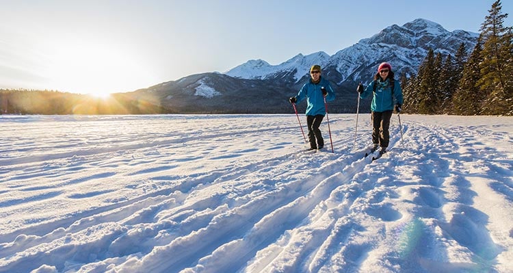 Two people cross country ski across a frozen lake below mountains.