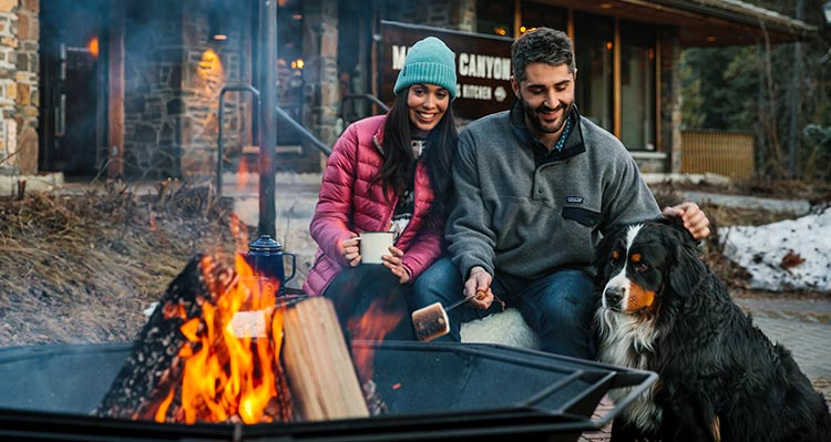 Two people sit with a dog at a campfire.