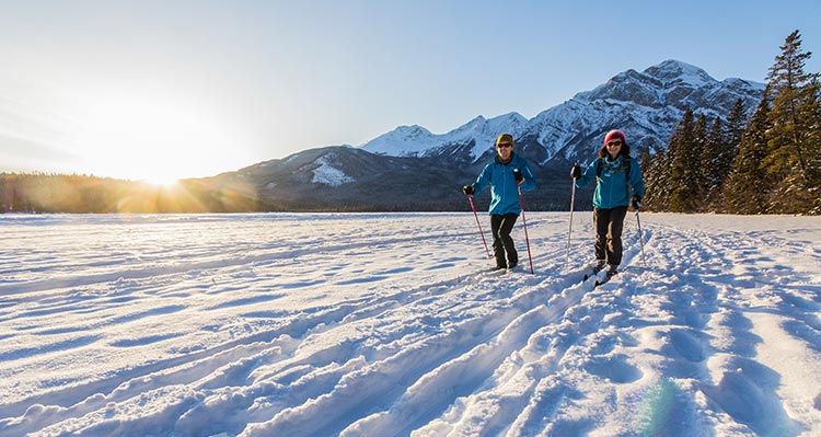 Two people cross country ski across a frozen lake below mountains.