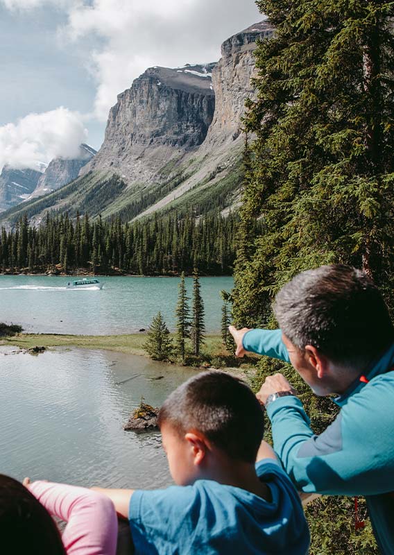 A family looks out at a small tree-covered peninsula on a lake below tall mountains.