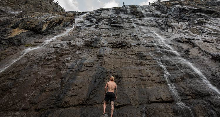 A person stands at the foot of a cliff with small waterfalls streaming down.