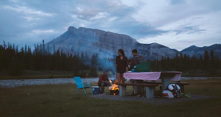 A group of friends cook food around a campfire at Two Jack Lake in the evening.