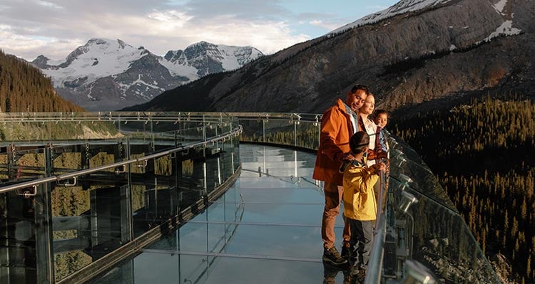 A family walks along a glass platform overlooking a forested valley.