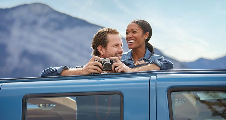 A couple peer out of the open rooftop of a bus, camera in hand