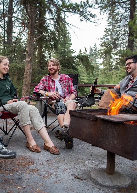 A group of friends sit around a firepit laughing and enjoying a beverage