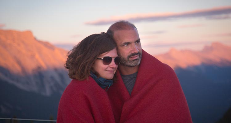 A couple huddle together with a blanket draped around them, gazing at the setting sun on the mountain top.