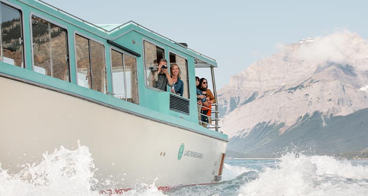 Passengers on a boat peer off the side to take photos, mountains behind