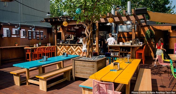 A patio with colourful tables and chairs, servers gather around the bar.