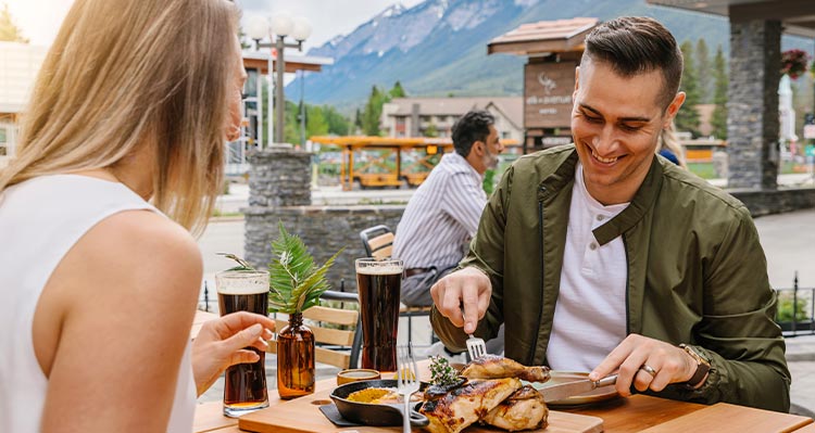 A couple sits on a patio, mountains behind, the man is cutting into his food.