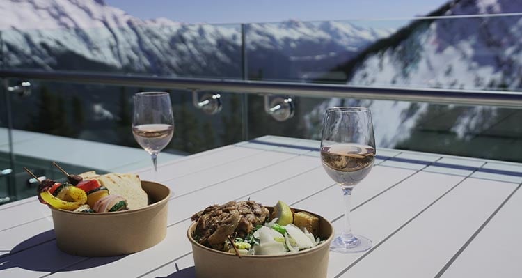 Two boxes of takeout food and two wine glasses sit on a patio table at the top of the mountain.