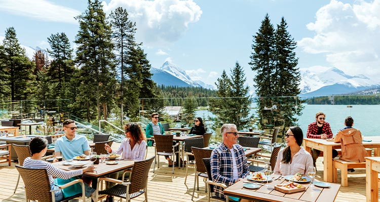 Groups of people sit at patios tables outside of a building, mountains in the background.