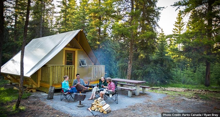 A group of friends sit around an outdoor fire at a campground with a tent fixture behind.