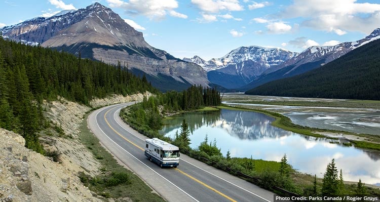 A bus drives down the road surrounded by trees, lake and a mountain.