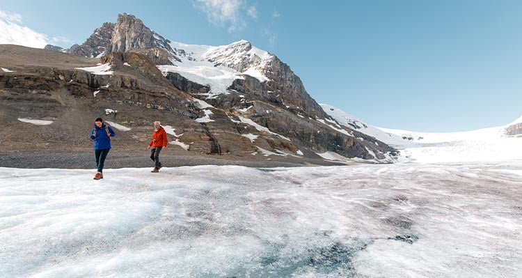 Two people walk along the Glacier ice. Mountains behind.