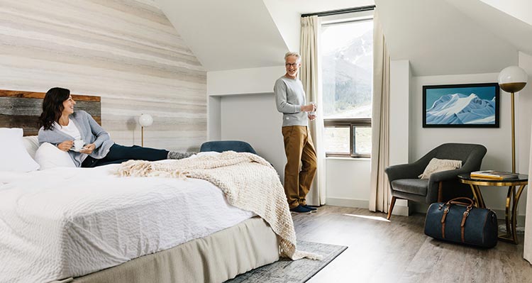 A couple enjoys the view from there room, the woman laying on the bed with a mug the man standing by the window.