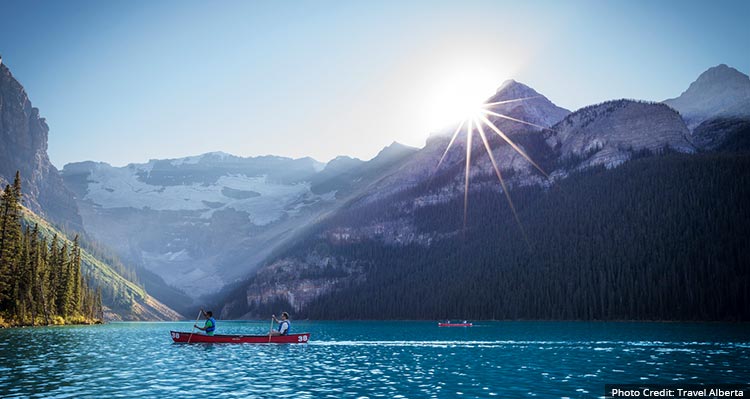 Two canoes with people paddle around the blue lake, sun is setting behind the mountains.
