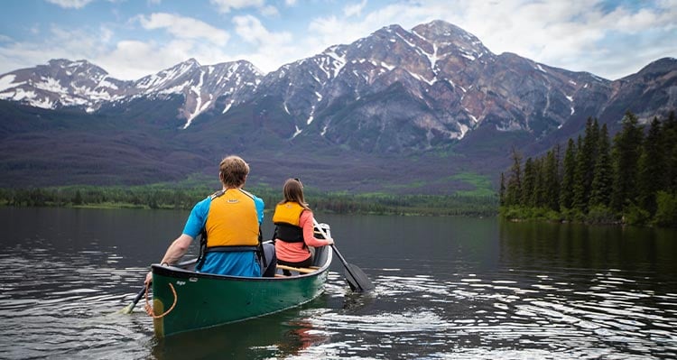 Two people sit in a canoe, paddling into the distance in a canoe, mountains in front of them.