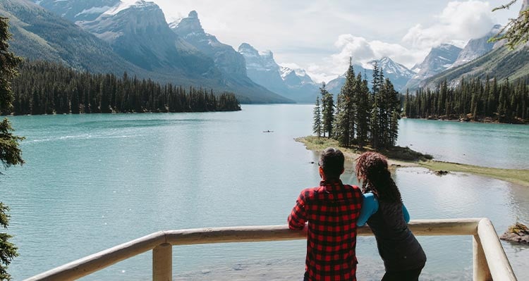 A couple leans over the railing looking out to the large lake surrounded by mountains.