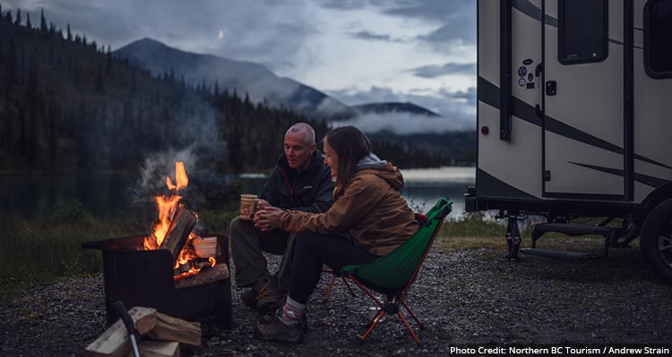 A couple sits at a campfire outside of a RV, mountain and lake behind.
