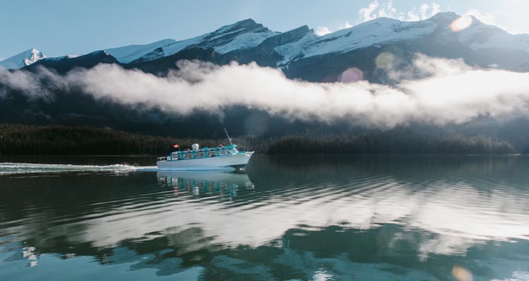 A blue boat cruises along calm water, mountains with snow behind.