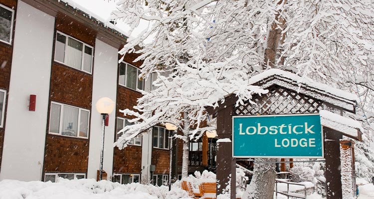 Snow covered hotel entrance surrounded by trees. Lobstick Lodge blue signage at the entrance.