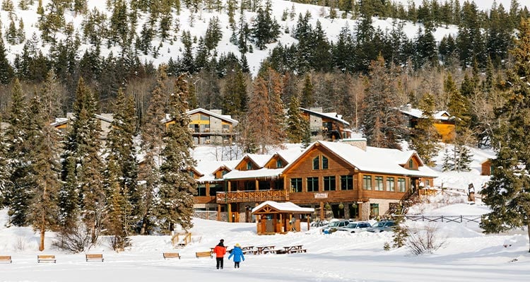 Two people walk across a snowy field towards a log cabin building.