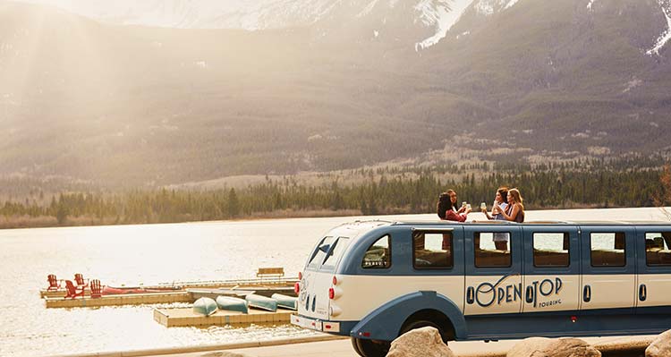 Women stand through the roof of the Open Top Touring vehicle, parked on the side of a lake.