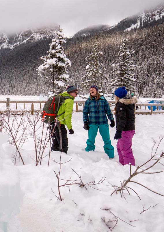 Three people smile and laugh in a winter wonderland scene next to an ice-carved Lake Louise sign.