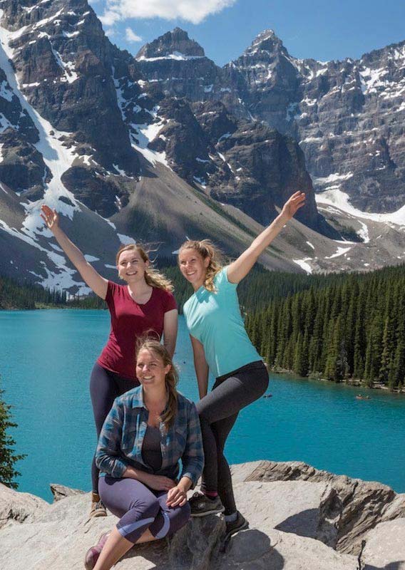 Three women pose for a photo above a blue lake surrounded by jagged mountain peaks