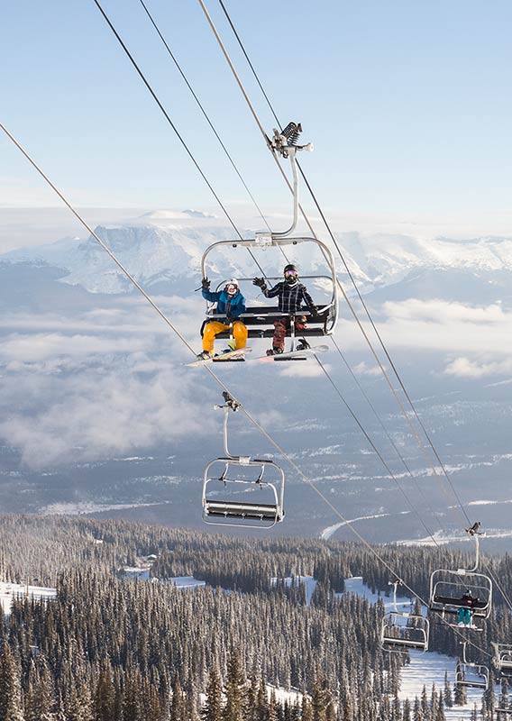 People wave from a chairlift above a forested, snow-covered mountainside.