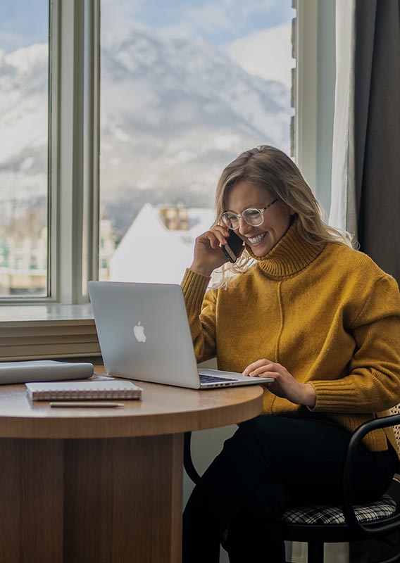 A woman works on a laptop in a hotel room at a wide window looking at mountain views.