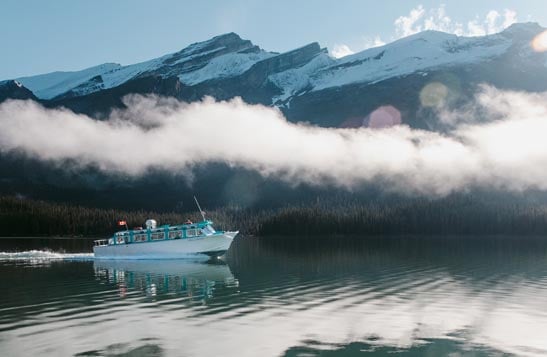 A boat cruises on a calm lake below low clouds and snow-covered mountains.