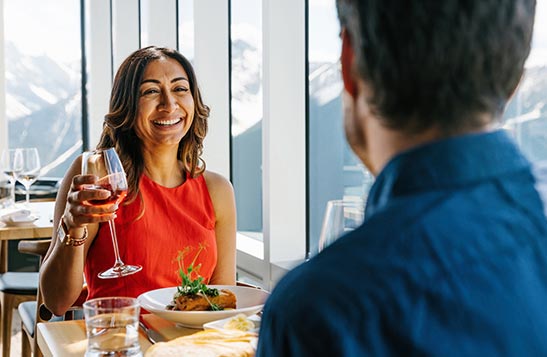 A woman raises a glass of wine at a dinner table against a sunny window.