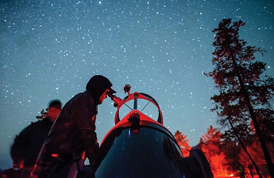 A man looks into a telescope outside with a starry sky above him